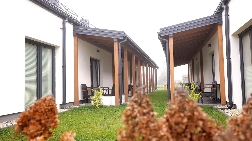 Zadaszone tarasy na drewnianych słupach między budynkami mieszkań wspomaganych, na pierwszym planie krzewy